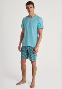 Lichtblauwe katoenen T-shirt gecombineerd met gestipte zwemshorts in teal, met geometrische vormen. Model staat blootsvoets met een neutrale uitdrukking.