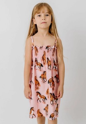 Junges Mädchen mit langen blonden Haaren, das ein ärmelloses rosa Kleid mit aufgedruckten galoppierenden braunen Pferden vor einem einfarbigen hellen Hintergrund trägt.