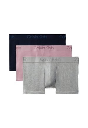 Uomo muscoloso in piedi a piedi nudi che indossa boxer neri Calvin Klein su uno sfondo grigio chiaro uniforme.