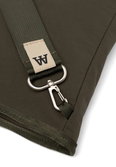 Pochette en tissu vert foncé à la surface texturée, dotée d'un clip en métal argenté et d'une sangle tissée avec une étiquette beige portant la mention "A".