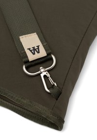 Un săculeț din țesătură de culoare verde închis, cu o suprafață texturată, având un clips metalic argintiu și o curea împletită cu un eticheta bej marcată cu „A.”