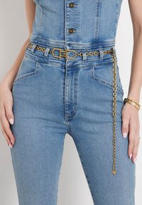 Zlatni lančani pojas s pravokutnom kopčom, nošen preko plavog traper kombinezona. Lanac sadrži okrugle karike, dodajući teksturu i detalj.