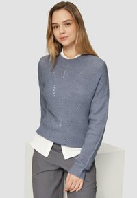 Pull en maille bleu clair avec un col rond, des poignets côtelés et des motifs en tricot ouverts, superposé à une chemise blanche à col.