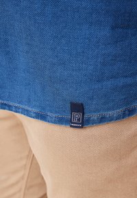 Blaues, strukturiertes Gewebe mit einem sichtbaren genähten Saum. Ein dunkelblaues Etikett mit dem Buchstaben "P" und dem Wort "Paddock's" ist nahe der unteren Kante angebracht.