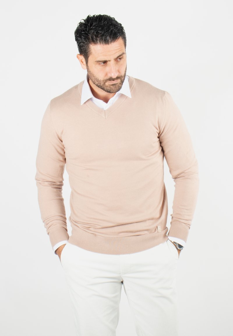 Pull en tricot beige avec un col en V, manches longues et poignets côtelés, associé à un pantalon blanc, comportant une étiquette de marque discrète à la taille.