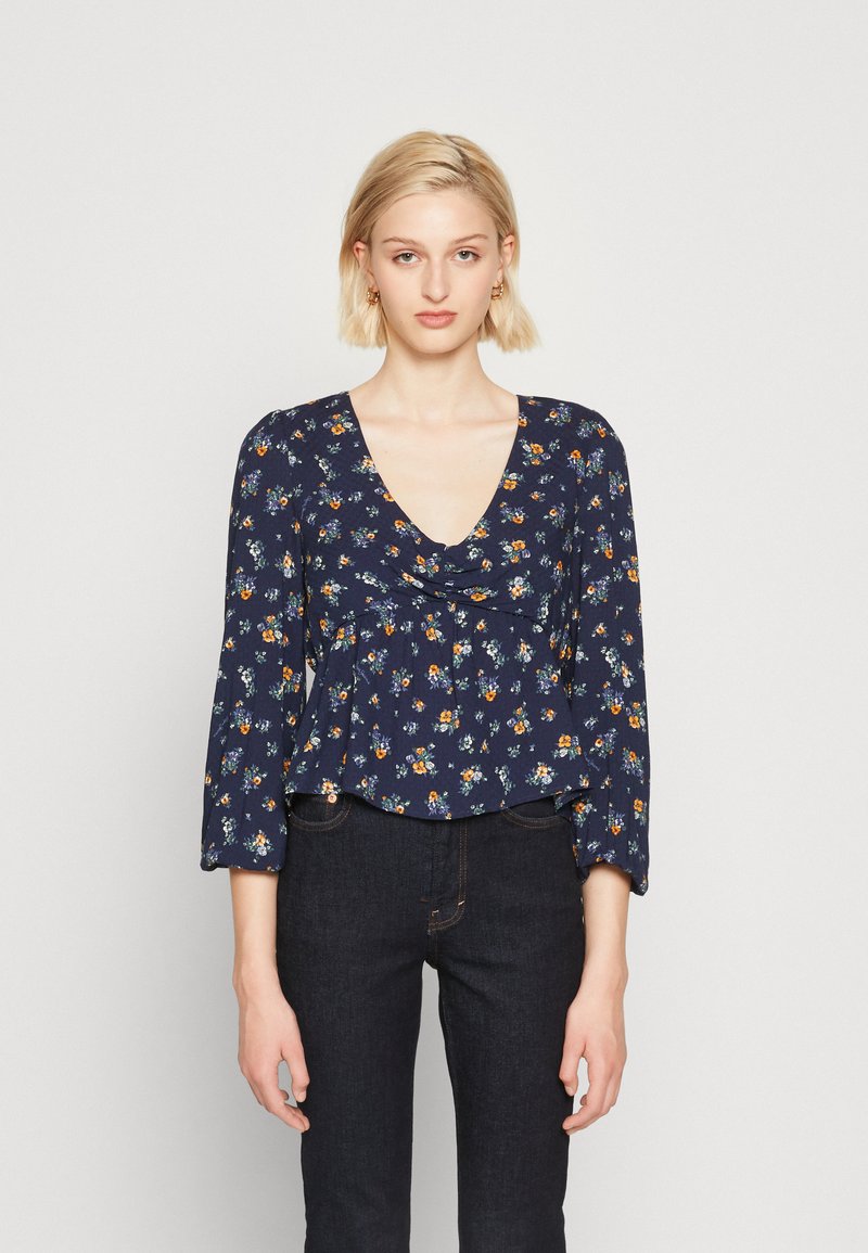 Donna bionda che indossa una blusa a maniche lunghe floreale blu navy con scollatura a V e jeans scuri a vita alta, in piedi davanti a uno sfondo neutro.