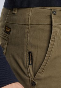 Khaki bomuldsbukser med en struktureret overflade, med en sort label og bronzefarvet knaplukning, delvist dækket af en marineblå ærme.