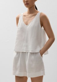 Top fără mâneci din in alb, cu decolteu în V, asortat cu pantaloni scurți lejeri, de același material. Ambele au o textură moale și o croială relaxată.