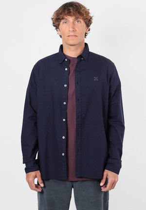 Homme aux cheveux bouclés portant une chemise bleu foncé boutonnée par-dessus un t-shirt bordeaux et un pantalon gris, debout devant un fond clair uni.