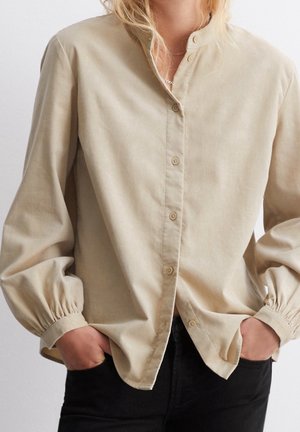 Personne portant une chemise beige à boutons avec poignets froncés et un pantalon noir, les mains dans les poches, sur un fond blanc uni.