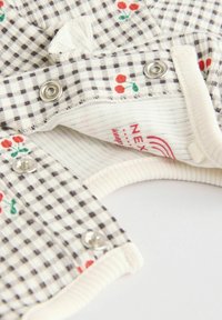 Kreminė ir pilka patikrinta medžiaga su raudonų vyšnių raštu. Yra metaliniai snapai ir raukšlėti kraštai, kuriuose matyti minkštas, tekstūruotas gamyba.