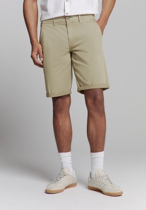 Khaki shorts gemaakt van lichtgewicht stof, met opgerolde boorden, een knoopsluiting en zijzakken. Draagt witte sokken en sneakers.