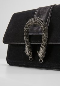 Borsa a tracolla in velluto nero con chiusura a pattina, caratterizzata da un manico metallico intrecciato a forma di teste di cavallo. Texture liscia e linee pulite.