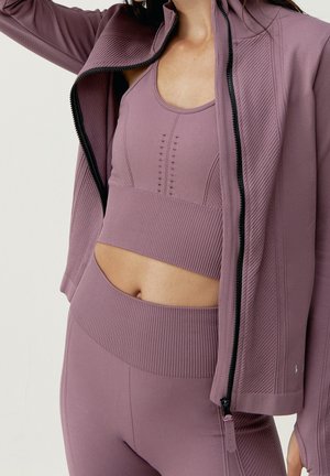 Frau trägt passenden, gerippten mauve Sport-BH, hoch taillierte Leggings und Jacke, mit teilweise geöffnetem Reißverschluss und erhobenem Arm.