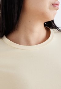 Top beige chiaro a maniche corte con scollatura arrotondata, texture liscia e tessuto morbido; primo piano della spalla e dell'area del collo.