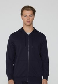 Námornícky kapucňový svetr na zip vyrobený z lehkého materiálu, s předním zipem, dvěma bočními kapsami a manžetovými rukávy.