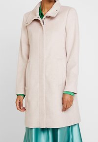 Cappotto beige chiaro fino al ginocchio con colletto alto sopra una blusa verde e una gonna di raso teal, indossato da una persona in piedi contro uno sfondo bianco.