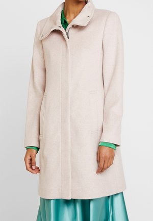 Cappotto beige chiaro fino al ginocchio con colletto alto sopra una blusa verde e una gonna di raso teal, indossato da una persona in piedi contro uno sfondo bianco.