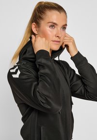 Schwarze Kapuzenjacke mit Reißverschluss aus glattem Material, mit hohem Kragen, seitlichen Taschen und weißen Logopunkten auf der linken Schulter.