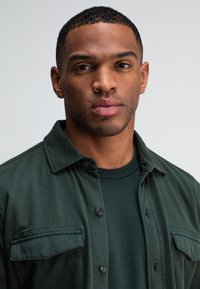 Grüne Button-Down-Bluse mit Kragen, versehen mit einer Brusttasche, über einem dunkelgrünen Rundhalsausschnitt-Shirt getragen. Glatter Stoff, strukturierte Passform.