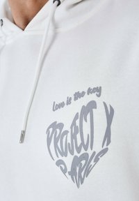 Sudadera blanca con capucha ajustable. Presenta texto gris en forma de corazón: "El amor es la clave" y "PROJECT X PARIS". Textura de tela suave.