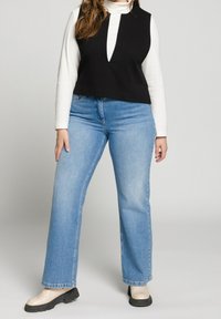 Svart ärmlös topp med V-ringning, lager på lager över en vit långärmad skjorta. Kombinerad med ljusblå utsvängda jeans och beiga skor.