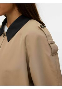 Beige jakke har en sort krave, skulderepauleter og runde knapper. Stoffet har en glat, let blank tekstur.