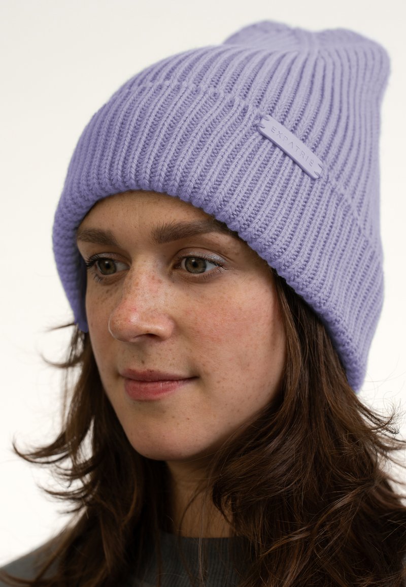 Lavendelfarbenes Strickmütze mit gerippter Textur, ausgestattet mit einem kleinen Etikett mit dem Markennamen. Die Mütze ist weich und lässig im Design.