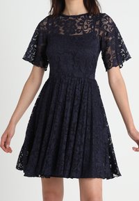 Marinblå spetsklänning med korta utsvängda ärmar, figurnära liv och utsvängd kjol. Har blommiga spetsmönster och fodrat liv för täckning.