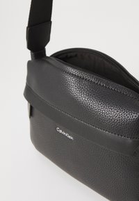 Borsa a tracolla nera in pelle sintetica con finitura testurizzata, dotata di chiusura con zip e design elegante. Logo visibile sul fronte.