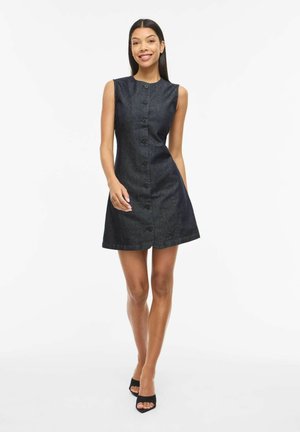 Uśmiechnięta kobieta w czarnej, bezrękawnikowej sukience zapinanej na guziki oraz czarnych butach na obcasie z odkrytymi palcami stoi na tle białego tła.