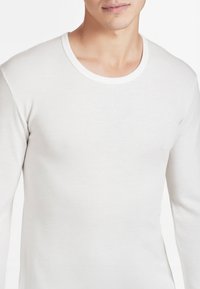 Maglietta a maniche lunghe e collo rotondo in tessuto beige chiaro. Superficie testurizzata con una finitura morbida e scollo bianco a contrasto. Design aderente.