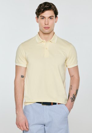 Młody mężczyzna z krótkimi brązowymi włosami nosi jasny żółty t-shirt polo i jasnoniebieskie spodnie, stoi z jedną ręką w kieszeni na jednolitym tle.