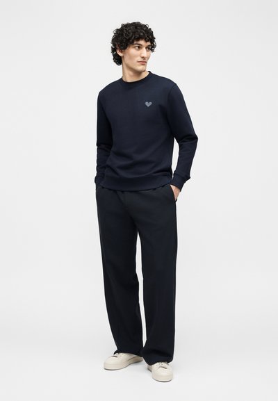 Άνδρας με σγουρά μαλλιά που φοράει ναυτικό μπλουζάκι με έμβλημα καρδιάς, μαύρο παντελόνι με φαρδιά μπατζάκια και λευκά αθλητικά παπούτσια, στέκεται με τα χέρια στις τσέπες.