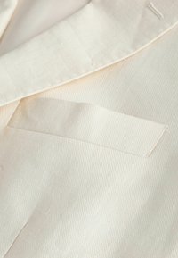 Gros plan sur un tissu de couleur crème avec une poche passepoilée sur mesure et des coutures visibles sur un matériau à texture lisse.