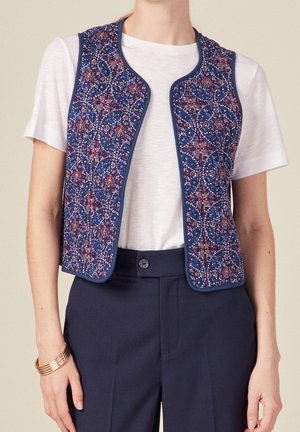 Vrouw draagt een aansluitend marineblauw vest met rood-wit bloemenpatroon over een wit shirt met korte mouwen en marineblauwe broek met hoge taille.