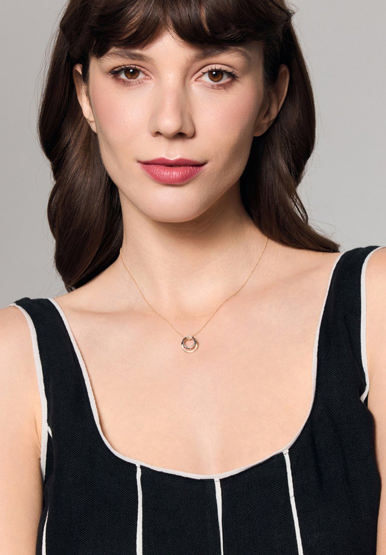 Złoty naszyjnik z okrągłym wisiorkiem, na którym znajdują się splecione pierścienie. Modelka ma na sobie czarną bluzkę w paski z kwadratowym dekoltem.