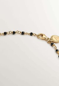 Cadena de oro con cuentas facetadas negras, presenta un cierre de langosta y una etiqueta redonda grabada con un patrón. Textura suave, diseño elegante.