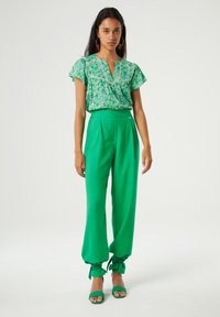 Groene bloes met korte gerimpelde mouwen, gecombineerd met groene high-waisted broeken. De broeken hebben verzamelde manchetten en een gladde textuur.