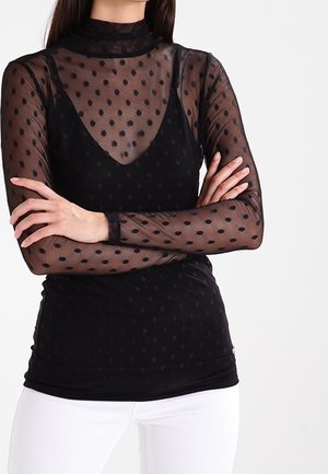 Femme portant un haut transparent noir à pois à manches longues superposé à un débardeur noir, assorti à un pantalon blanc, bras croisés.