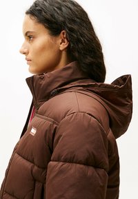 Giacca puffer isolante marrone con cappuccio, dotata di zip frontale e cuciture verticali a baffle. Texture morbida e logo del brand sul petto.