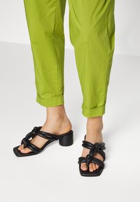 Zwarte hoge sandalen met geknoopte bandjes, vierkante neuzen en een chunky hak. Gecombineerd met groene cropped broek die enkeldetails laat zien.