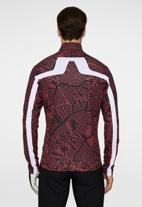 Långärmad sporttröja med ett svart och röd vågigt mönster, med vita accenter på axlarna och ärmarna. Sitter tight.