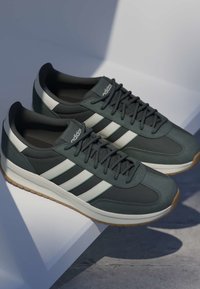Paire de baskets Adidas vert foncé avec des bandes blanches, des semelles beiges et des lacets noirs, placée sur une surface blanche inclinée avec des ombres.