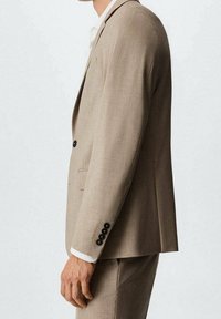Giacca di costume beige su misura con finitura testurizzata, design a un petto, bottoni neri sulle maniche e una camicia bianca sotto.