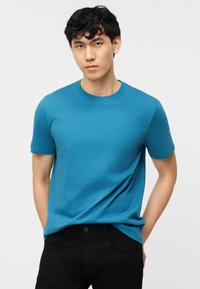 Νεαρός άντρας με μαύρα κυματιστά μαλλιά, που φοράει απλό μπλε κοντομάνικο μπλουζάκι και μαύρο παντελόνι, στέκεται με τα χέρια στις τσέπες.