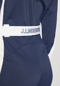 Tumedast vormi kohandav ülikond valgete detailidega. Omab valget vööd, millel on tekst "J. LINDEBERG". Sile tekstuur, kaasaegne disain.