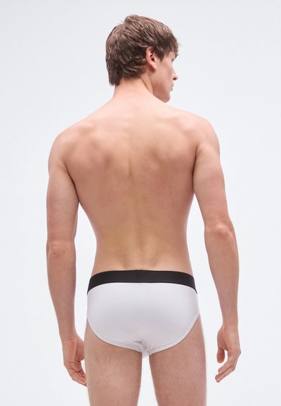 Braguitas blancas de algodón con una banda elástica negra en la cintura, vistas desde atrás. Textura suave y diseño ajustado que realza la forma del cuerpo.