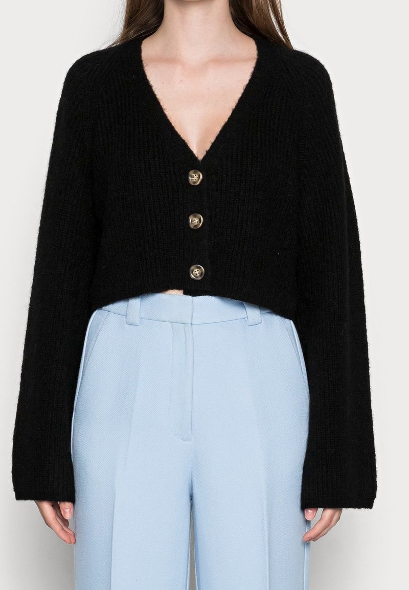 Černý zkrácený kardigan ve strukturovaném úpletu, s hlubokým výstřihem do V a třemi zlatými knoflíky vpředu. Nosit s světle modrými kalhotami s vysokým pasem.