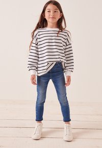 Csíkos, hosszú ujjú ing fehér és tengerészkék színben, laza fazonnal és ráncolt mandzsettával, kék slim fit farmernadrággal és fehér sportcipővel párosítva.